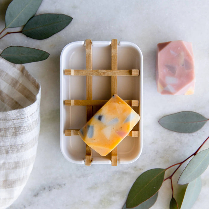 Orange soap bar on a wooden soap dish with green leaves and a textured container in the background.