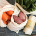 Mesh produce bags with carrots, sweet potatoes, and a roll of organic cotton produce bags on a dark surface.