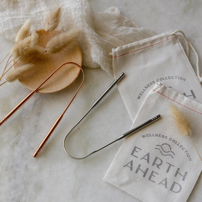 Metal U-shaped  tongue scrapers on a marble surface with 'Earth Ahead' branded bags.