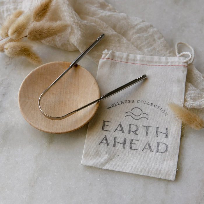 Metallic U-shaped tongue scraper on a wooden plate with a 'Earth Ahead' branded bag on a light background